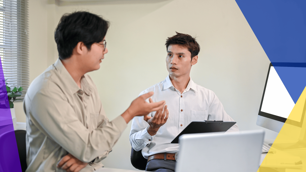 Two men in an office having a discussion, one gesturing with his hands, laptops on the desk beside them.