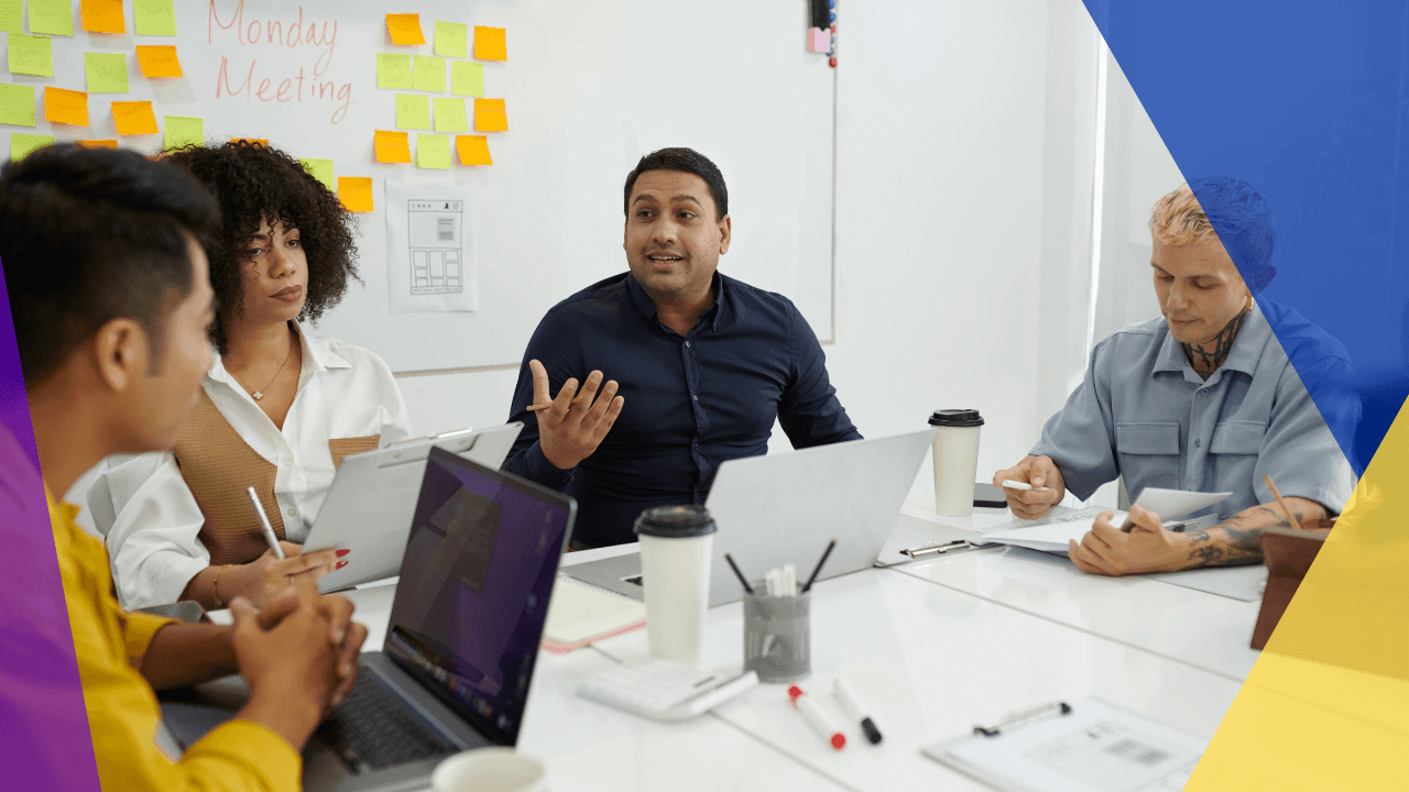 Man in navy shirt gesturing at a Monday Meeting whiteboard covered in sticky notes while three colleagues listen at a table with laptops.