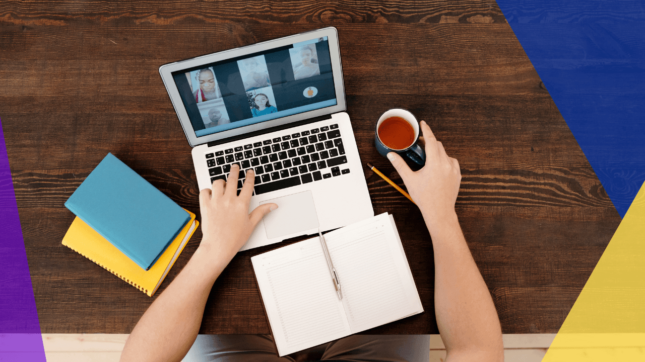 Overhead view of hands on a laptop showing a video call grid, holding a mug of tea beside a notebook on a wooden table, purple accents.