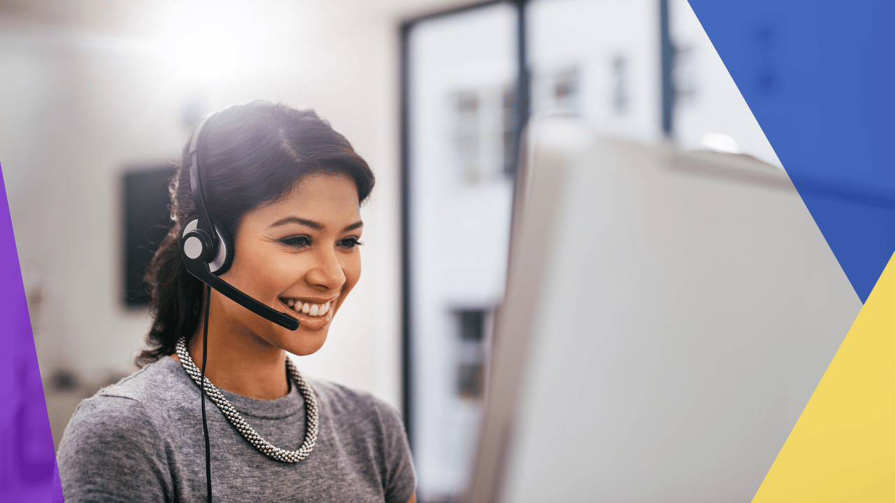 Smiling call center agent in a headset and grey top looking at a computer monitor, bright blurred office behind.