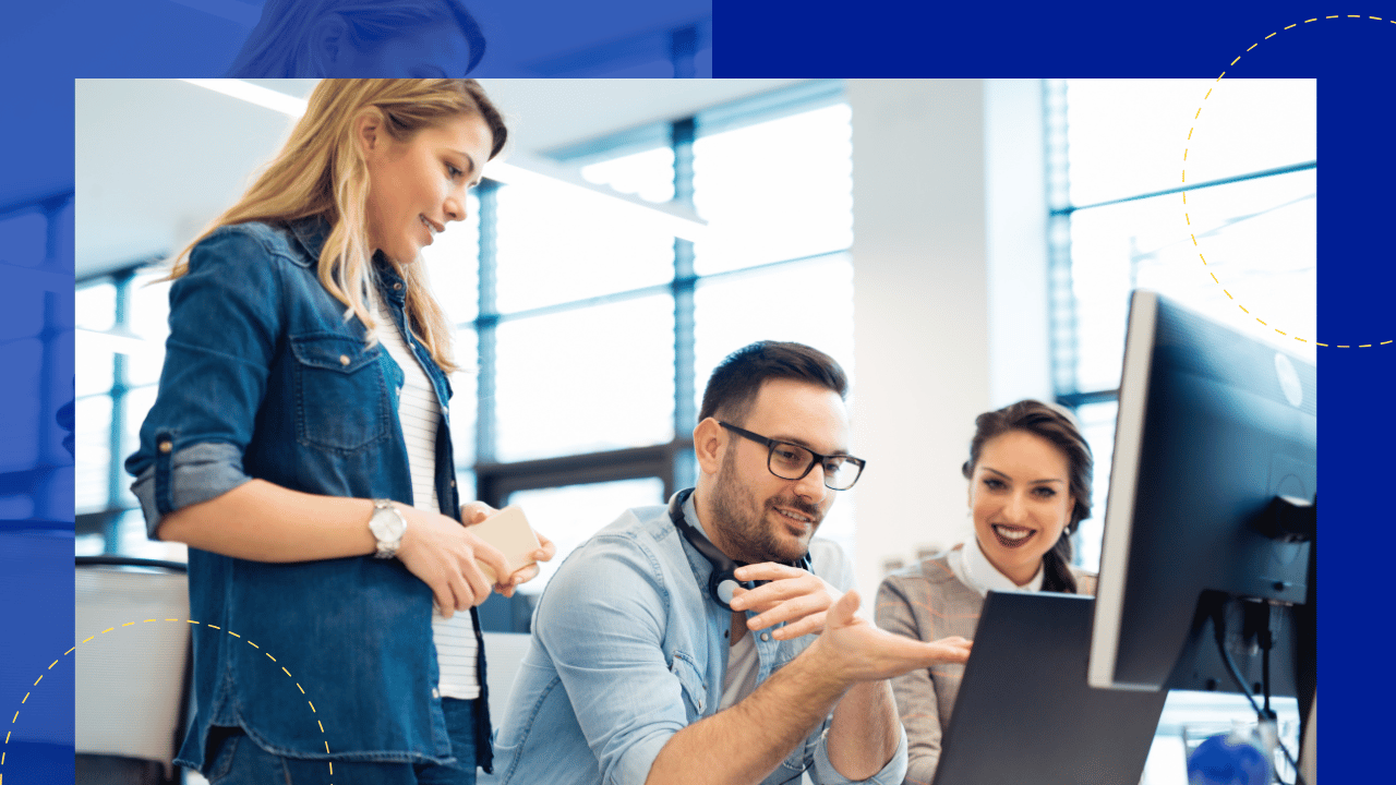 Man in glasses explaining something at a desktop monitor to two smiling female colleagues in a bright office.