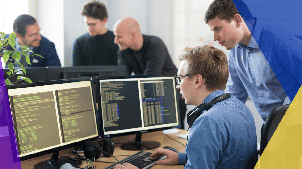 Developer in glasses and headphones at two monitors of code while a colleague watches, three teammates talking behind.