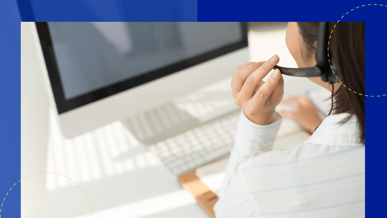 Call center agent adjusting a headset microphone at a white desk with an iMac and keyboard, bright window light.