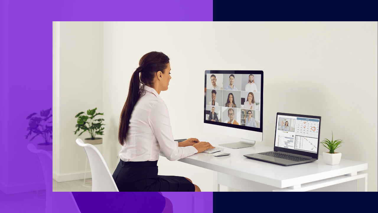 Woman at a white desk on a video call with a grid of nine colleagues on a monitor, laptop showing charts beside her.