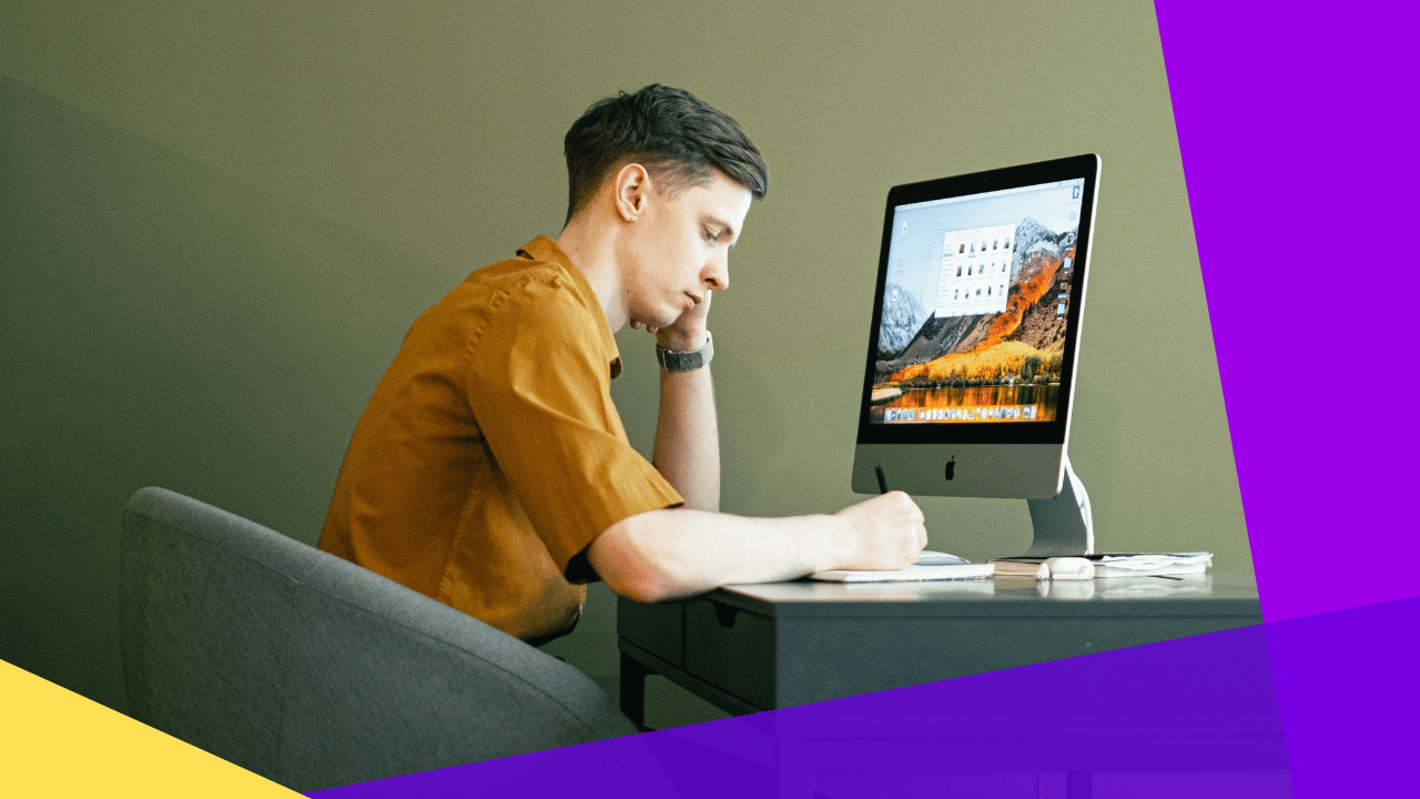 Young man in a mustard shirt writing at a desk in front of an iMac, chin resting on his hand, plain olive wall behind.