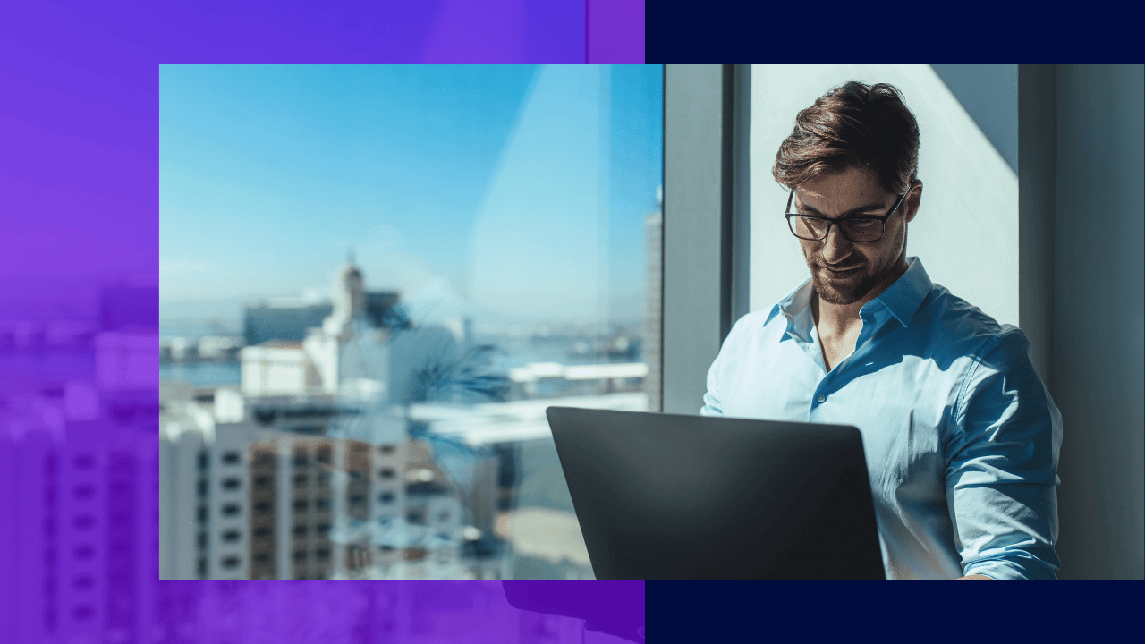 Man in glasses and a light blue shirt working on a laptop beside a high-rise window overlooking a city harbour, bright daylight.