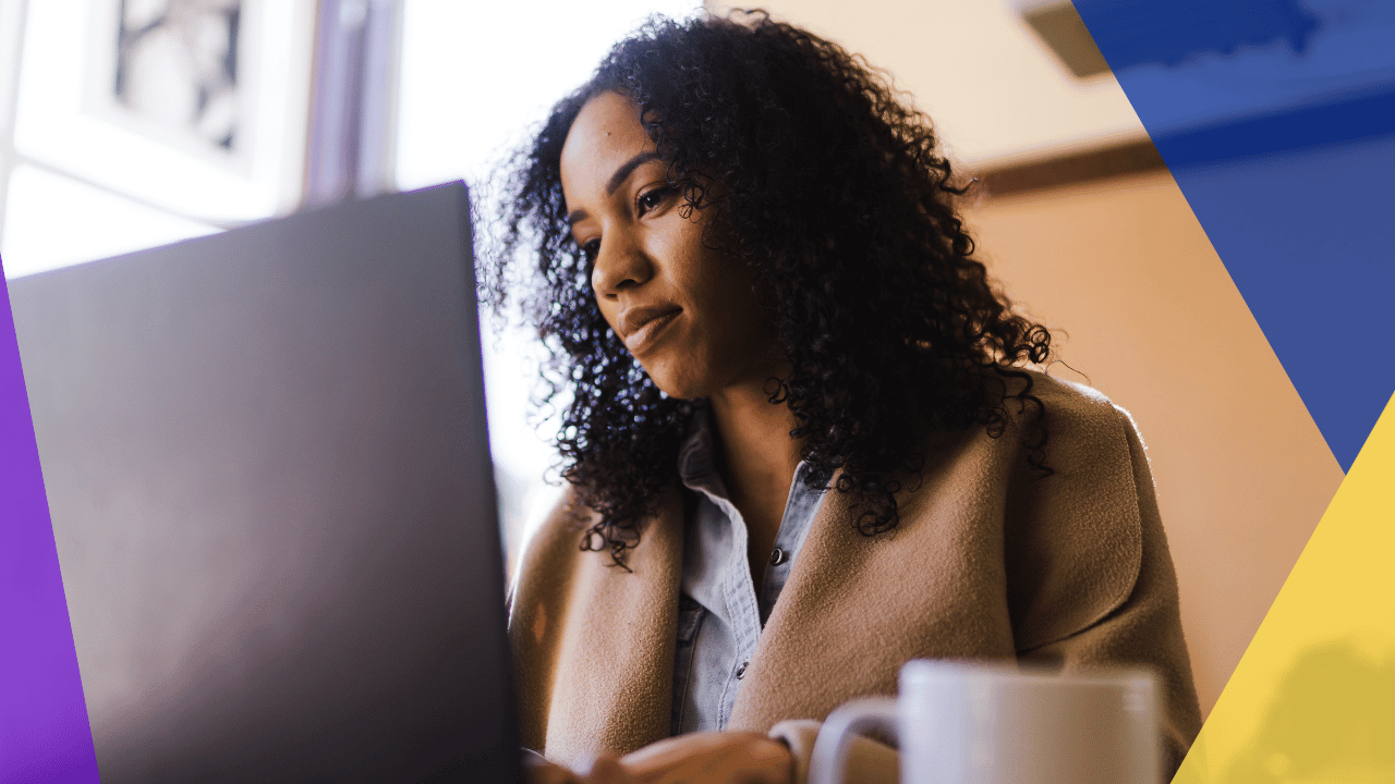 Woman with curly hair reading a laptop screen at a table beside a white mug, warm window light from behind.